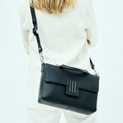 Skinnveske i Svart med Gulldetaljer. Hippi Grace Sotogrande Midi er en mellomstor veske til dame i ekte skinn. Skinnvesken kommer med et elegant håndtak og en avtagbar og regulerbar crossbody stropp. Vesken har et klassisk, funksjonelt og tidløst design som overlever alle trender. Skinnvesken har en klipp lås i front for å lukke vesken. Veske dame. Sort veske. Svart veske dame. jobbveske dame. hverdagsveske dame. Norsk design. Crossbody Veske dame. Crossbodyveske dame.  Modellbilde med vesken [wv]